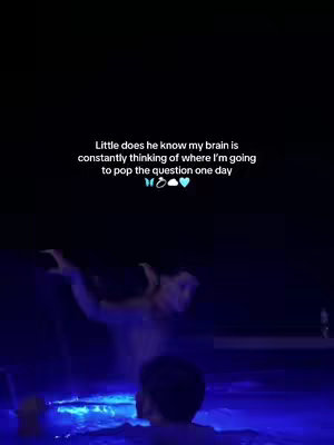 An overhead shot shows two people in a pool at night with a text overlay. The water is illuminated with blue lights, reflecting on the figures and the surrounding area. The text states "Little does he know my brain is constantly thinking of where I'm going to pop the question one day".