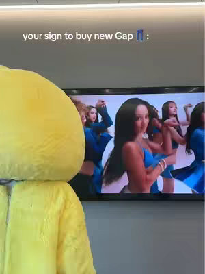 A person wearing a large, yellow costume resembling a duck stands in front of a television. The television displays a colorful music video with female performers in blue outfits. The costume covers the person's body and head, obscuring their identity. The top of the frame is visible, showing the hook text.