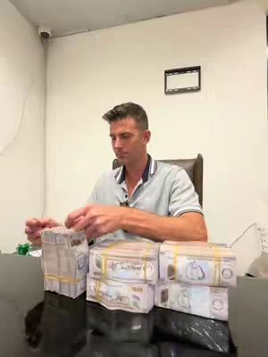A man in a light gray polo shirt sits at a dark surface surrounded by stacks of bundled, banded cash in a bright, well-lit room. A security camera can be seen in the upper left of the frame. The man looks down, appearing to interact with the money or objects nearby.