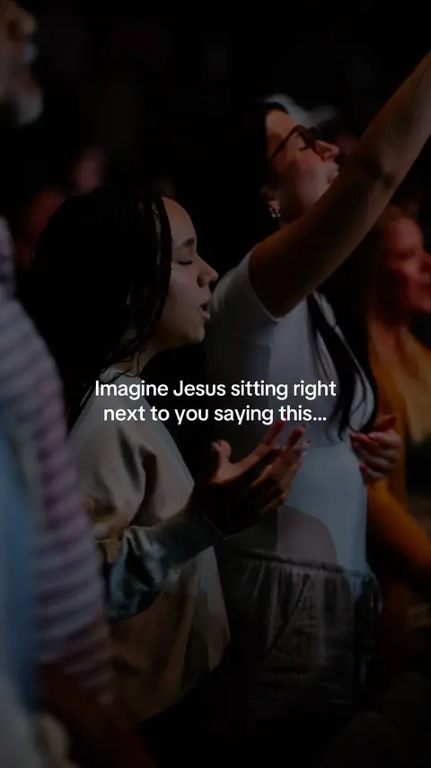 The image captures a moment in a dimly lit setting, likely a worship service or gathering, with several individuals in the foreground. The central focus is on three people: two women and a partial view of what appears to be a man on the left. One woman is central, eyes closed, and her hands raised in a gesture of worship or praise, wearing a light-colored top. The other woman on the right, partially visible, in also having her hands raised. The figures appear to be positioned in front of a wider group, suggested by the blurred shapes in the background.