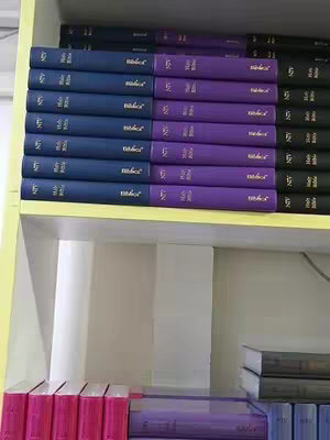 The image shows several stacks of books. The books are primarily blue and purple, arranged on a shelf. Some of the covers are visible, with gold lettering. The scene is well-lit with a neutral shelf background.