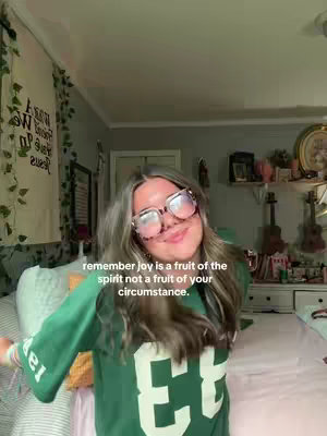 A young woman with glasses and long hair smiles in a bed room setting, wearing a green jersey. The background includes a partially visible bed, decorative items on a shelf, and a drawn-on banner. The overall lighting is natural.