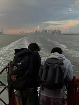 Two people are standing on the back of the Staten Island Ferry, facing away from the camera. The sky is a mix of gray and warm colors, and the New York City skyline is visible in the distance. Water churns behind the ferry, and the scene captures a tranquil moment on the water.