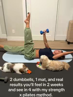 A woman is lying on a mat indoors, performing a pilates exercise. She has one leg extended upwards and is holding a dumbbell in her other hand above her. Several dogs are resting on one side of her body. The setting appears to be a home gym.