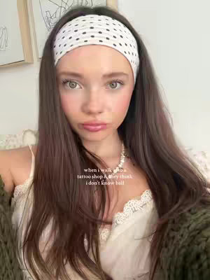 A young woman with long brown hair and a white polka dot headband is the focus. She's in front of a light background with some art pieces visible. She's wearing a white top and looks like she's in her room. Text overlay is added to the bottom of the image. She has a neutral expression.