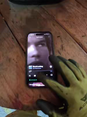 A person's gloved hand holds a smartphone, displaying a close-up image of a face reflected from the phone screen. The phone is placed on a wooden surface. The hand is wearing a gardening glove. The display shows the music app interface and song "Good Looking". The foreground is dominated by the phone and hand, with the background featuring a wooden surface with warm tones.