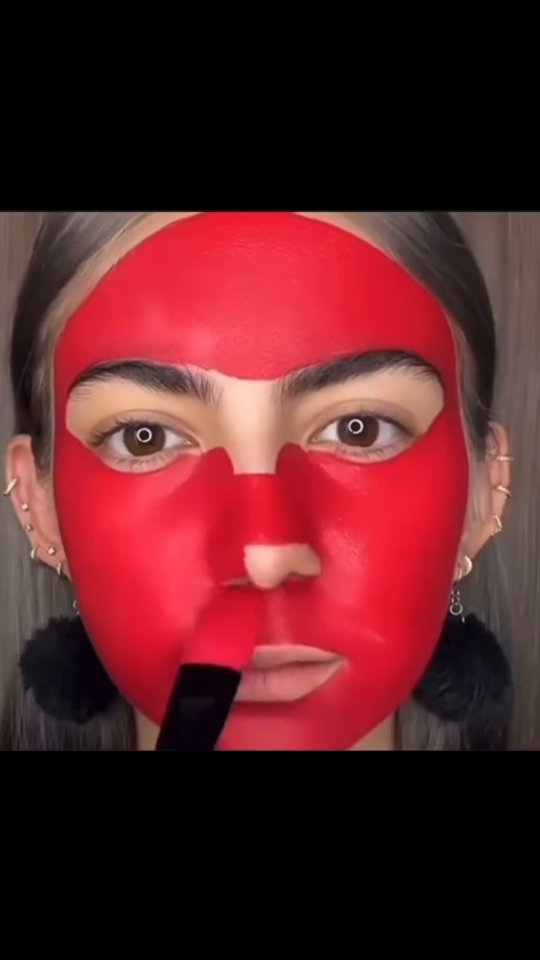 The cover image is a close-up of a person's face, primarily covered in bright red makeup. The makeup covers the entire forehead, cheeks, chin, and extends onto the nose, with defined areas for the eyes. A black makeup brush is being held by the person and is applying the red makeup to the lips. The person has light-colored skin and visible eyebrows, along with fair eyelashes and visible dark eye sockets. There are earrings, a series of piercings, two black fluff ball earrings, dark long eyelashes and a portion of dark hair at the top of the head.