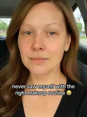 A woman with long, wavy brown hair is shown in a medium shot. She is in front of a blurred background, possibly inside of a car. The woman's expression is neutral, conveying a sense of anticipation or self-reflection. She has minimal makeup on. The text overlay at the bottom of the image says, "never saw myself with the right makeup routine" with a face palm emoji.