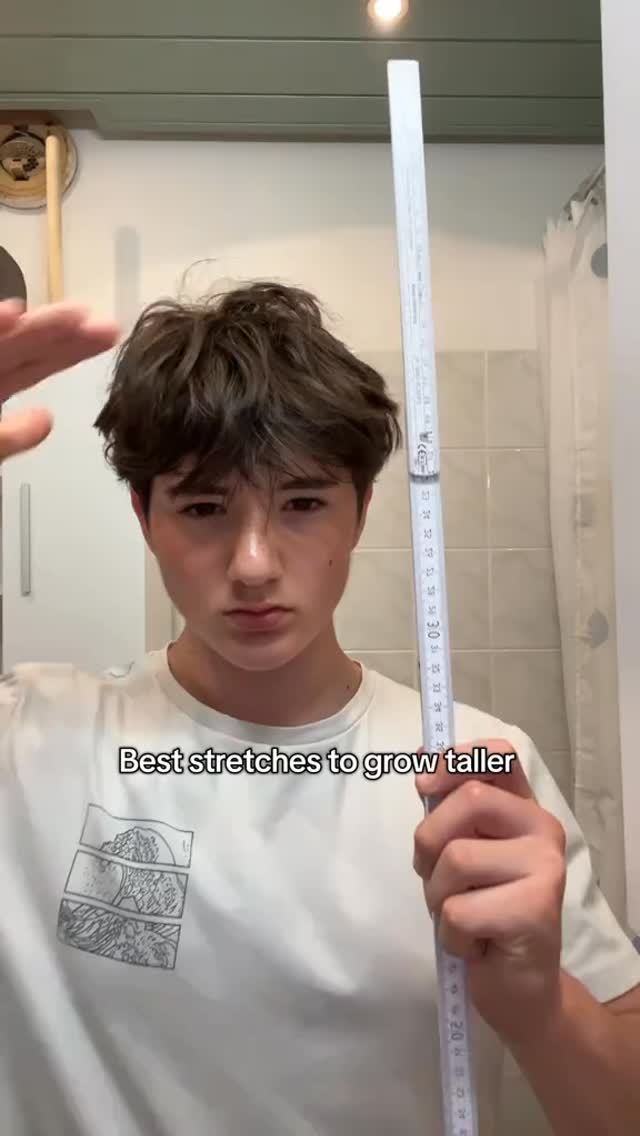 A young man stands indoors holding a measuring stick, looking towards the camera. Text overlayed reads 'Best stretches to grow taller'. The background is a bathroom setting.