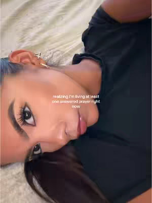 A selfie-style shot of a woman lying on a bed. She is positioned with her head near the bottom and leaning to the side, looking directly at the camera. The background appears to be a bed with light-colored, textured bedding. She has long dark hair, styled with a side part. She has noticeable makeup, particularly around her eyes and lips, which draws attention. The lighting seems to be soft, possibly natural or diffused, which illuminates her face without harsh shadows. A text overlay indicates that she might be talking about her faith or answered prayers.