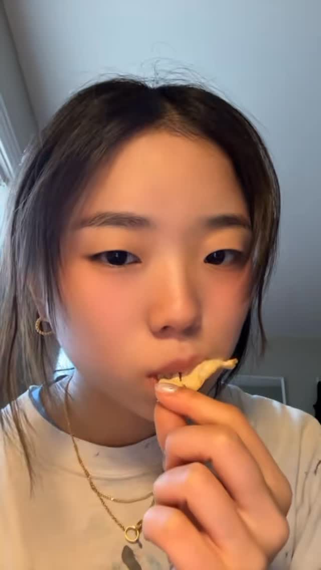 A close-up shot of a young woman eating a chip, with her face taking up most of the frame. She is wearing a simple white t-shirt and a delicate gold necklace. Her eyes are dark, and she has rosy cheeks. Part of her hand is visible, holding the chip as she brings it towards her mouth. The background is subtly blurred, suggesting an interior setting.