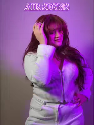 A woman is seen in a vertical shot with a purple background and a text overlay. The woman has long, dark hair and is wearing a light-colored, zip-up top. She is touching her head with one hand. The overlay text reads "AIR SIGNS" in a bright, neon-style font.