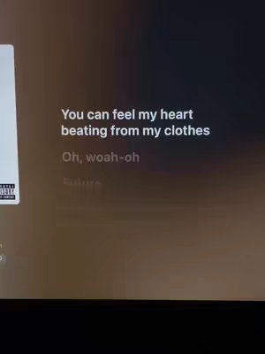 A screenshot from a screen showing a song's lyrics. The lyrics are white text over a blurred, dark background.