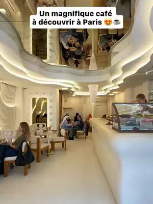 An interior shot of a cafe with a white, modern design. The ceiling has a wavy, cloud-like structure with embedded lighting. There are tables and chairs, and people are seated and conversing. The background includes architectural details and reflections. A display case is visible, and a person serves behind a counter. The colors are bright and airy.