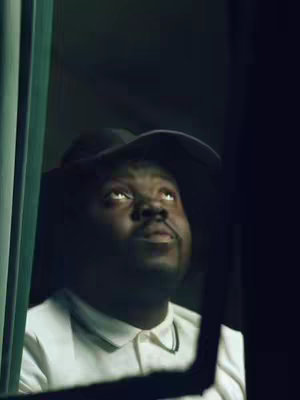 The image shows a man with dark skin looking upwards with his eyes. He is wearing a white collared shirt and a black hat. The background is out of focus and dark.