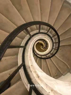 An overhead shot of a spiral staircase, viewed from the top looking down. The steps are a beige color, with a dark, curved handrail outlining the inner curve of the spiral. The overall composition emphasizes the circular and descending nature of the staircase.