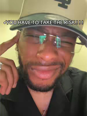 A close-up shot of a Black man wearing a baseball cap and glasses. The man is touching his temple with his index finger and has a slight frown. Overlay text saying, 'YOU HAVE TO TAKE THE RISK!!!' is at the top of the image.