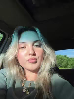 A selfie taken inside a car. The woman is looking towards the camera. She has blonde, wavy hair and is wearing a necklace. Outside the window, green trees and blue sky are partially visible.