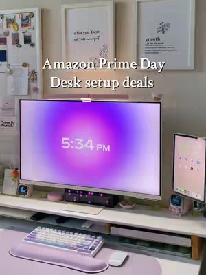 The image features a well-lit, modern desk setup with a large monitor displaying a purple and white gradient background and the time. A white keyboard, mouse, and desk accessories are in the foreground, with a tablet on a stand to the right. The background includes framed artwork against a light-colored wall, with the text 'Amazon Prime Day desk setup deals' overlaid at the top.