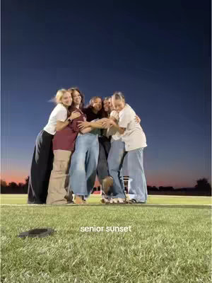 A group of six young women in casual attire, arms wrapped around each other in a tight embrace. They are standing on a well-kept grass field with a sign that reads \"senior sunset\" against a backdrop of a deep blue sky and a sliver of orange and pink at the horizon. The lighting suggests it's dusk.