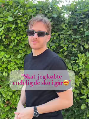 A medium shot features a young man standing in front of a lush green hedge. He wears sunglasses, a black t-shirt, and a watch. Text overlay is present near the bottom, in a speech bubble shape.