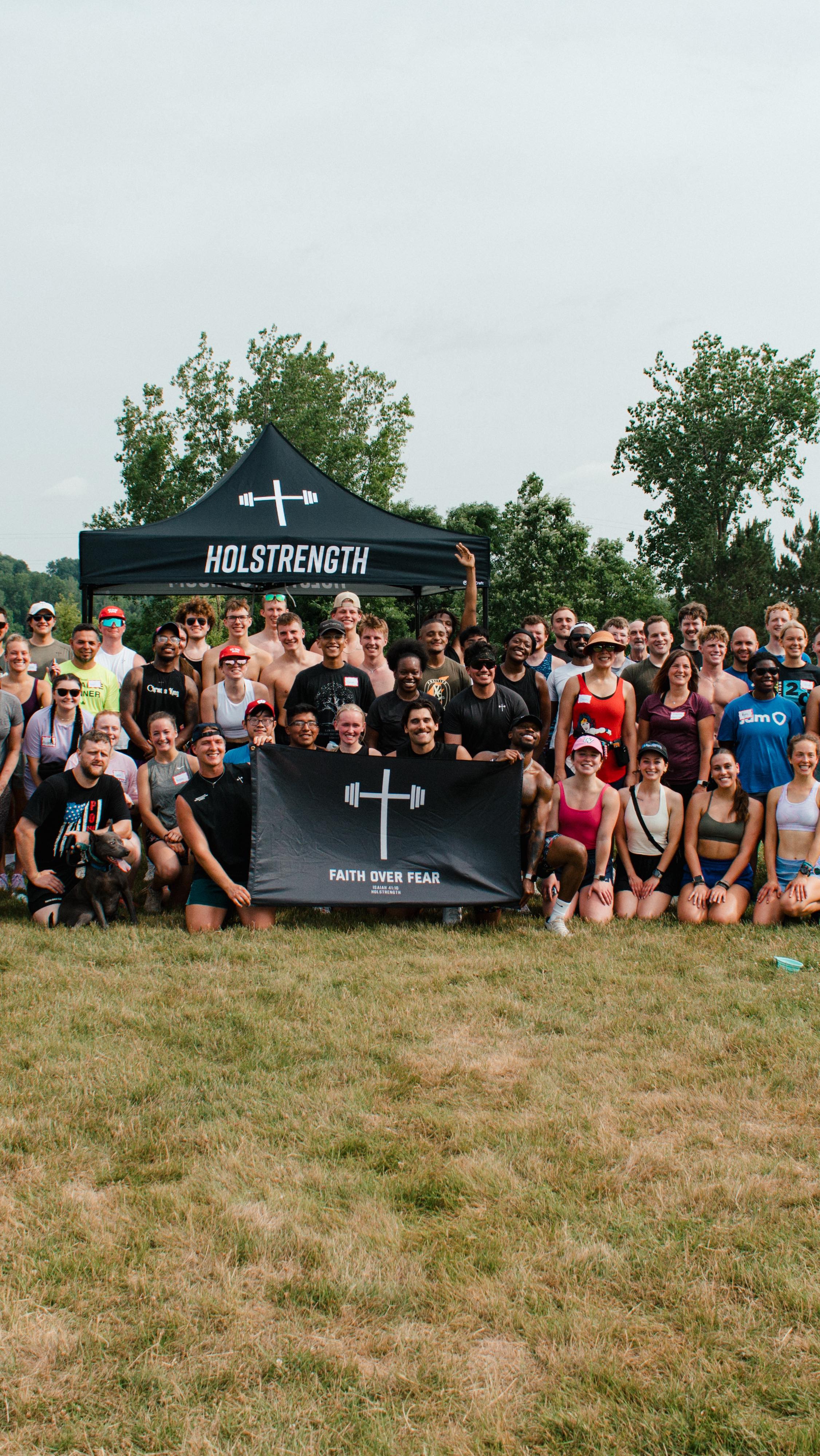 A group photo of a large gathering of diverse people on a grassy field. A black tent labeled 'HOLSTRENGTH' is in the background. A banner with a cross and the words 'FAITH OVER FEAR' is held by some of the people in the front.