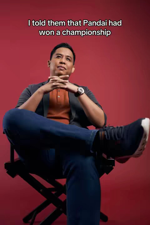 A man sits in a director's chair with legs crossed, hands clasped, looking up. The background is a solid red. Text overlay at the top.