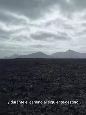The image shows a wide shot of a volcanic landscape under a cloudy sky. In the foreground, the terrain appears dark and textured, likely composed of volcanic rock. The middle ground features a line of low-lying hills or mountains, and the background is dominated by higher peaks fading into the overcast sky. The overall color palette is muted, with shades of gray and brown creating a somber yet captivating atmosphere.