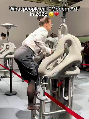 The image shows a person interacting with a sculpture in what appears to be an art exhibit. The person, likely a woman, is wearing a white shirt and dark skirt, and is interacting with the structure. The background is blurred, suggesting a crowded space, likely an art gallery or museum. The image is well-lit and the colors are natural.