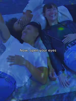 A group of three people are lying down, appearing to be looking up at something. A person in the foreground is wearing a white t-shirt, while another has an arm extended toward the camera and is partially obscured. The person closest to the camera is a man and he is smiling. The lighting is blue, creating a cool ambiance.