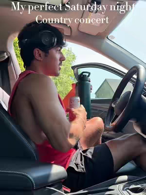 A man with short dark hair is seated in the driver's seat of a car with the door open. He's shirtless, wearing a red tank top, black shorts, and headphones. He has a dark beverage in his hand. The background features a sunny outdoor scene with a green field and a house.