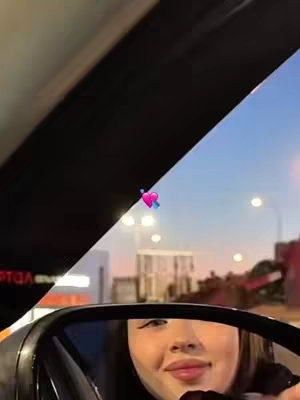 The cover image shows a woman in the reflection of a car's side mirror. The scene is set at night with city lights and a colorful sky visible in the background. The woman is smiling softly, the heart emoji on a wing adds a fun touch.