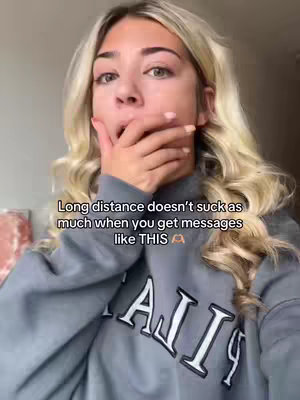 A young woman with long wavy blonde hair is covering her mouth with her hand, eyes wide, expressing surprise. She is wearing a grey sweatshirt with white text, positioned approximately in the center of the image. Text is overlaid at the bottom of the image.