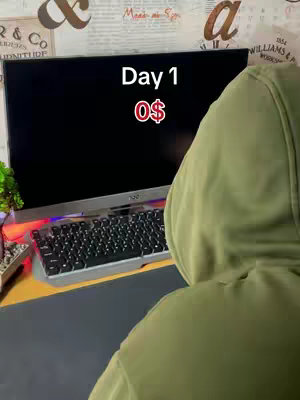 A shot from behind a person wearing an olive green hoodie, positioned in front of a computer setup. The screen displays a black screen, with white text reading 'Day 1' above the number '$0', colored in red. A keyboard and small plant can be seen on the desk in front of the screen.  The background shows a textured wall with decor.