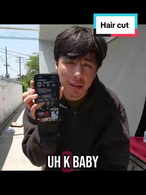 A man holds up his phone displaying an app or social media profile. The man is looking directly at the camera, and text overlays say "Hair Cut" and "UH K BABY".