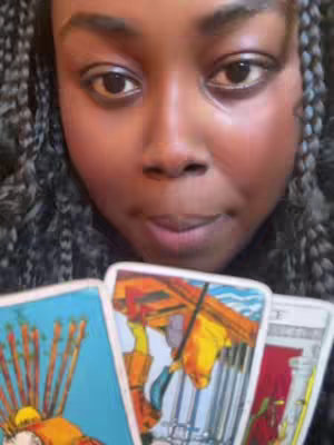 A close-up shot features a person's face with dark skin, centered in the frame, looking slightly downward. The person is holding up three tarot cards in the foreground, partially obscuring the lower part of their face. The background consists of their braided hair.