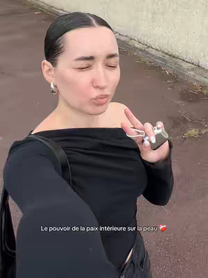 A woman with slicked-back dark hair pouts her lips, closes her eyes, and makes a peace sign with her right hand. She's wearing silver hoop earrings and a black off-the-shoulder top. In her right hand, she holds a car key remote. The shot is taken from a slightly low angle, capturing her from the chest up, against a blurred outdoor background with a road and a wall.