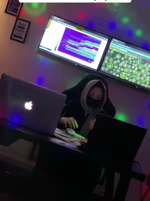 A person wearing a hood and a mask sits at a desk with two laptops and a stack of money. Two screens are mounted on the wall behind them.
