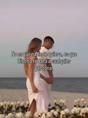 A couple, a man wearing glasses and a woman with long hair and wearing a white dress, are standing on a beach. The sea and sky are visible in the background. The woman is turned away from the camera, and the man is facing towards the camera. White flowers are placed in the foreground.