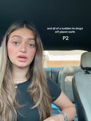 A woman is sitting in the driver's seat of a car, looking directly at the camera and talking. The car's interior is visible, and text overlays appear on screen. Her hair is long, and she's wearing a black shirt and various bracelets. The background includes the car's overhead and a window.