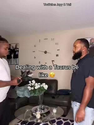Two men stand in a living room setting. One man on the left is holding a phone and looking at the other. The man on the right, with a beard is looking at him. In the foreground is a round glass-top coffee table with flowers and candles. A clock is on the wall. The text overlay says, "Dealing with a Taurus be like 🥺".