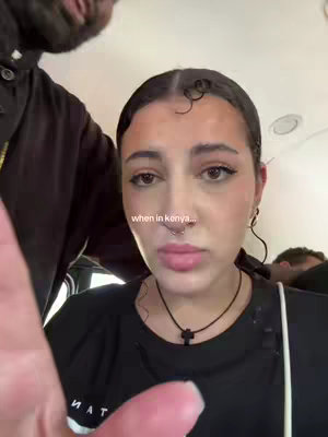 A selfie style video cover image shows a person in a black shirt from the chest up. The person is pale-skinned with thin eyebrows and is looking towards the camera. The person's hair is pulled back. Text that reads, "when in kenya..." is overlayed on the image in the lower center of the frame. In the top left corner there is a portion of a person's face and beard.