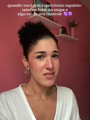 A woman with dark hair styled in a bun is looking at the camera with a serious expression. The background is a solid pink wall with white shelves. Text overlays at the top of the image describe astrology themes..