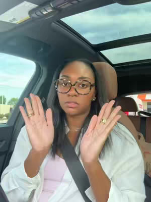 The cover image shows a woman inside a car, looking directly at the camera with a slight expression of surprise or amusement. She has dark hair, is wearing glasses, and has her hands up with palms forward as though showing something or making a gesture. Her seatbelt is visible, and the inside of the car is visible, with a sunroof overhead and some greenery visible outside the windshield. Her attire includes a light pink top and a white top over it.