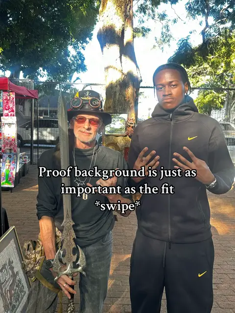 Two men stand outside in front of a shop with displays. One man wears a steampunk-style hat, with sunglasses on, and holds a large sword. The other man wears a black hoodie. The hook text overlays the image.