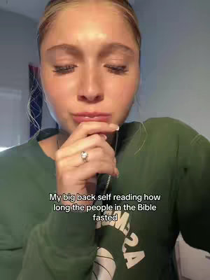 A selfie-style shot featuring a young woman with blonde hair and closed eyes. Her right hand is raised to her mouth, with a ring visible on her finger. She is wearing a dark green sweatshirt. The background is blurred, but appears to be indoors. Text is overlaid at the bottom, reading 'My big back self reading how long the people in the Bible fasted'.