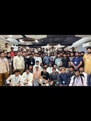 A photograph taken indoors featuring a large group of men, likely students or participants, posing together. They are casually dressed and have name tags, gathering together in the room. The lighting appears to be overhead, and the background is plain, with neutral-colored walls, indicating a professional or educational setting.