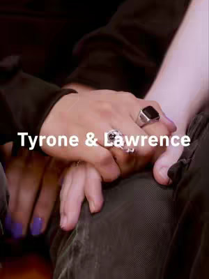 The image shows two hands holding each other, with the hands of two people of different genders. A hand with purple nail polish is resting on top of another hand. The hands feature rings, including a large black square ring on the top hand. The text overlay reads, 'Tyrone & Lawrence'.