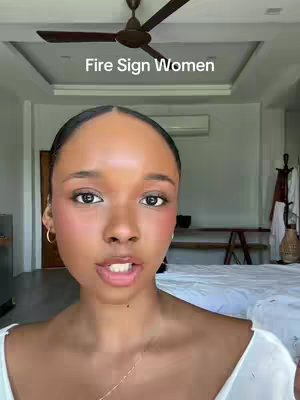 A woman with dark hair and light makeup is looking directly at the camera. Above her head, text reads "Fire Sign Women". The background is a room with a ceiling fan, a bed, and a door.