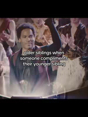 The image shows a scene from The Hunger Games. The character in the foreground, a Black man with short, styled hair, is slightly smiling and raising one arm with a clenched fist. Text overlays the image, reading 'older siblings when someone compliments their younger sibling'. The background is filled with blurry figures of people, suggesting a crowd or audience. The color palette is muted with shades of brown, beige, and black.